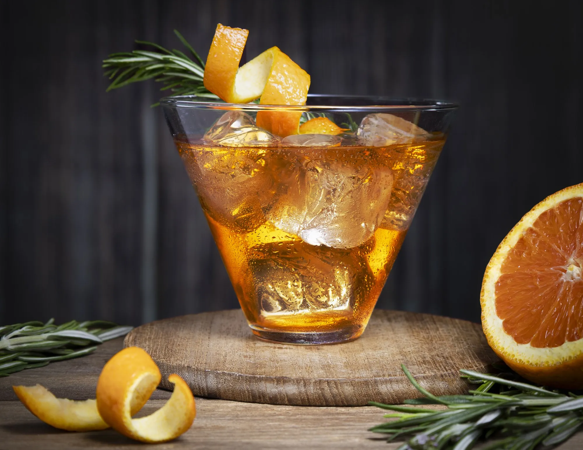 An amber cocktail with orange and rosemary glows against a dark tabletop.