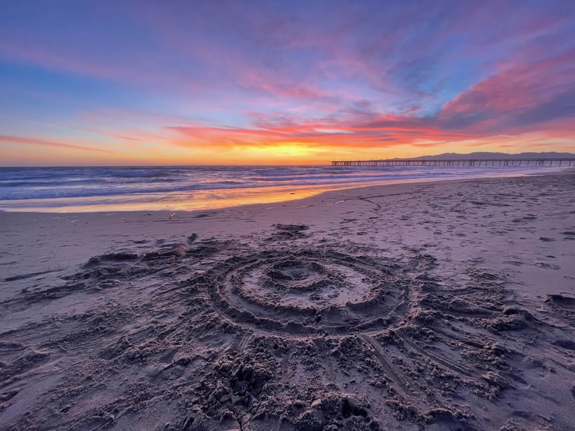 A circular labyrinth is traced into the beach beneath a colorful evening sky.