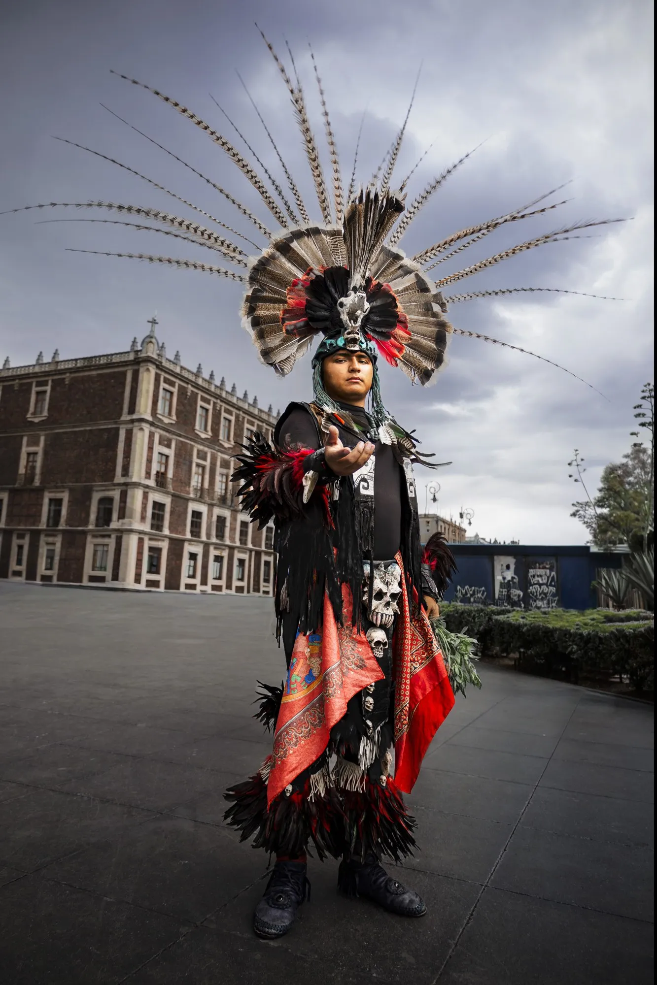 A costumed dancer in a tall red-and-black headdress stands in a city square.