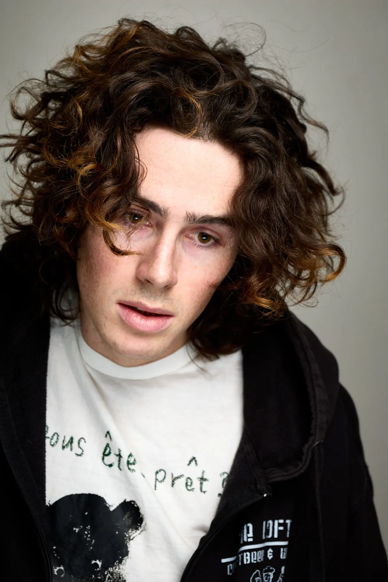 A curly-haired young man raises both hands to his head in a low-key portrait.
