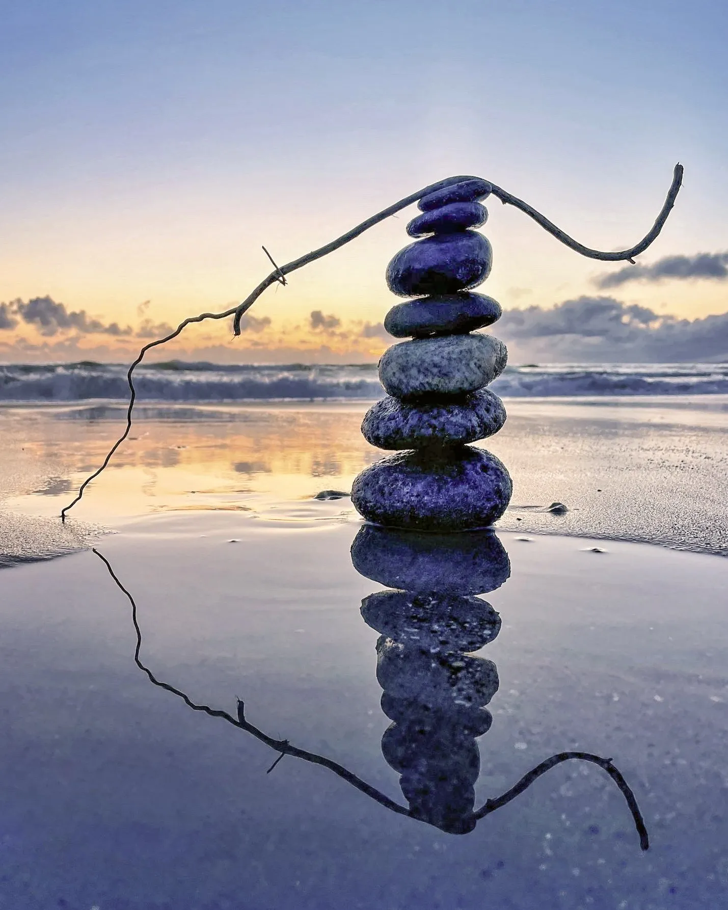 A curved driftwood branch arches over a balanced stone tower and its reflection.