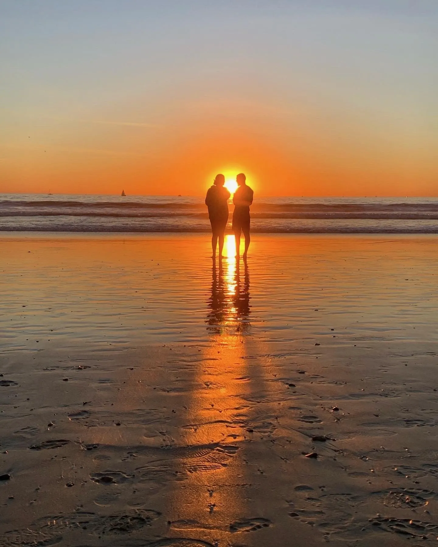 A figure stands in the bright reflection of the setting sun on wet sand.