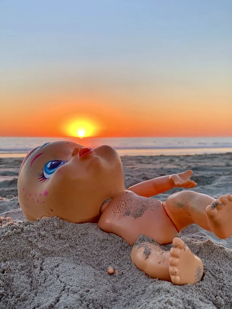 A toy baby doll on the beach appears to blow the setting sun like a bubble