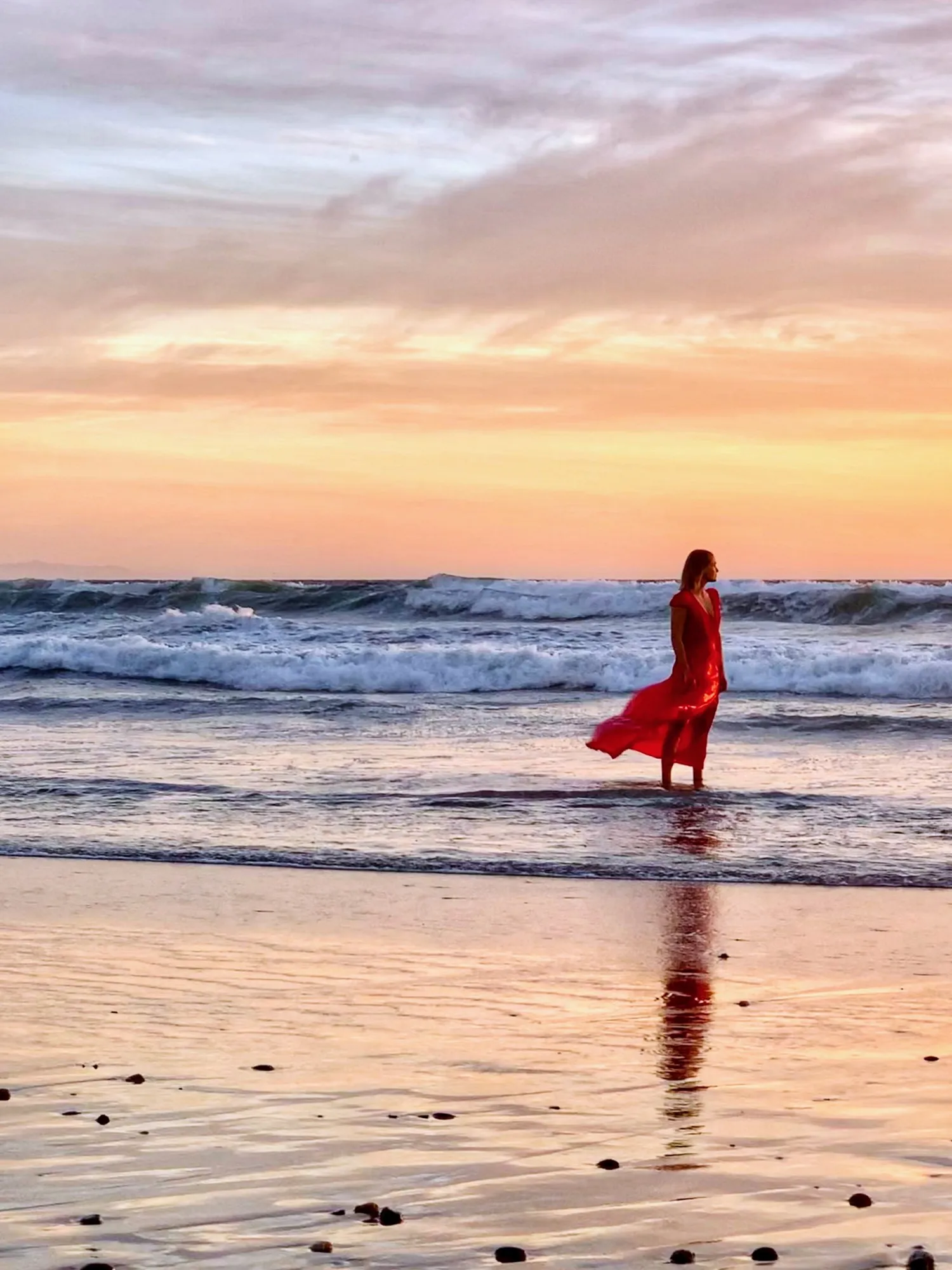 A woman in a flowing red dress standing in the surf beneath a peach sky