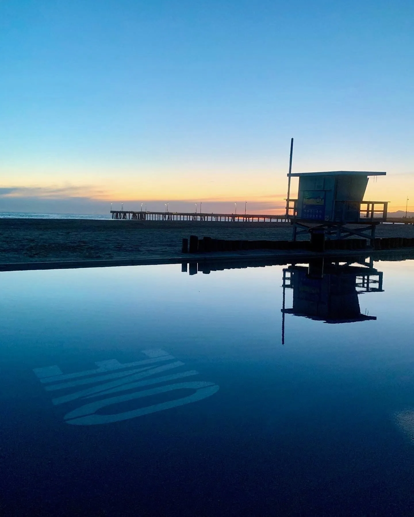 A lifeguard tower reflected in still water beside the pier at dusk