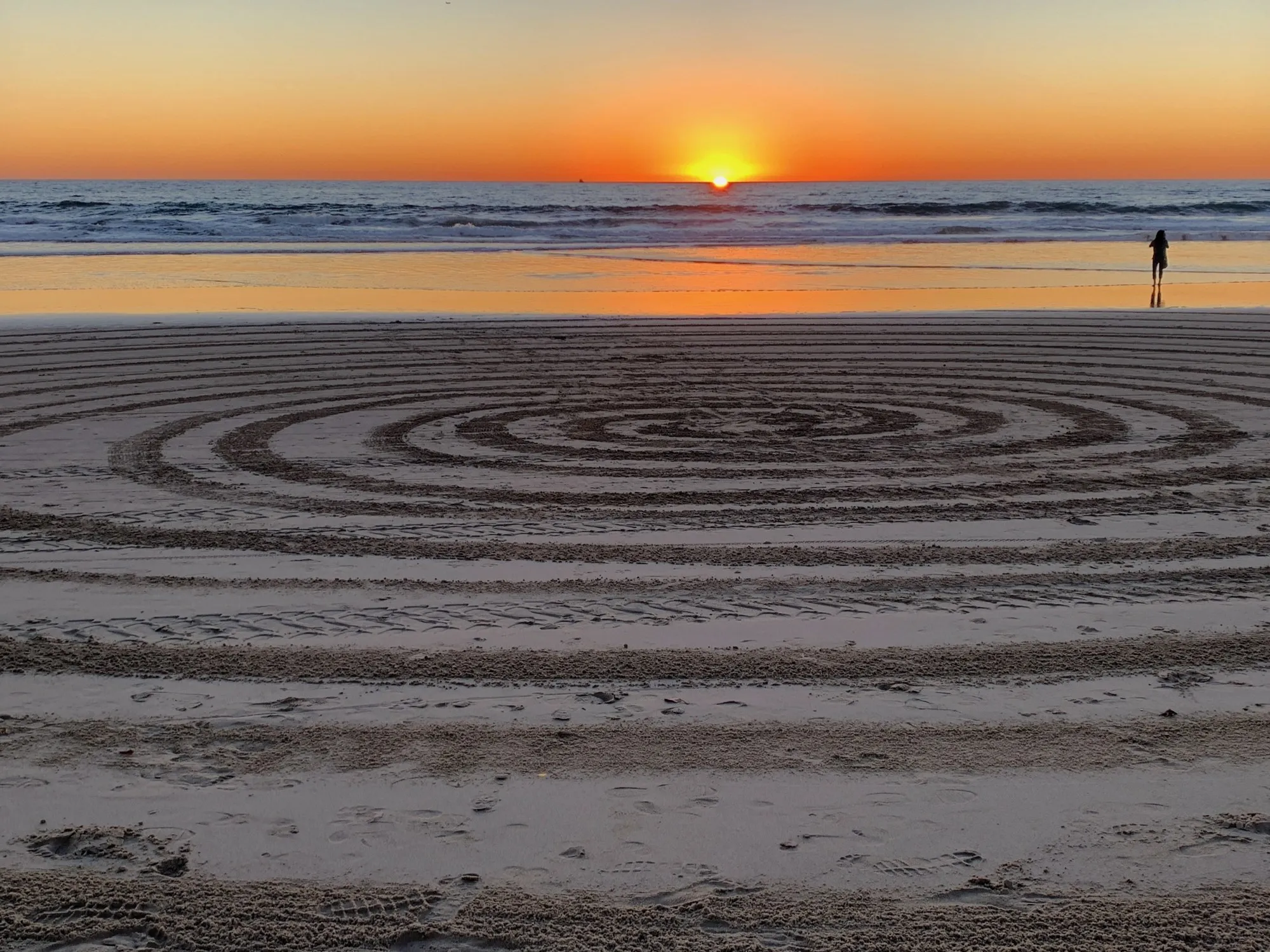 A spiral drawn in beach sand with a lone figure and the setting sun beyond