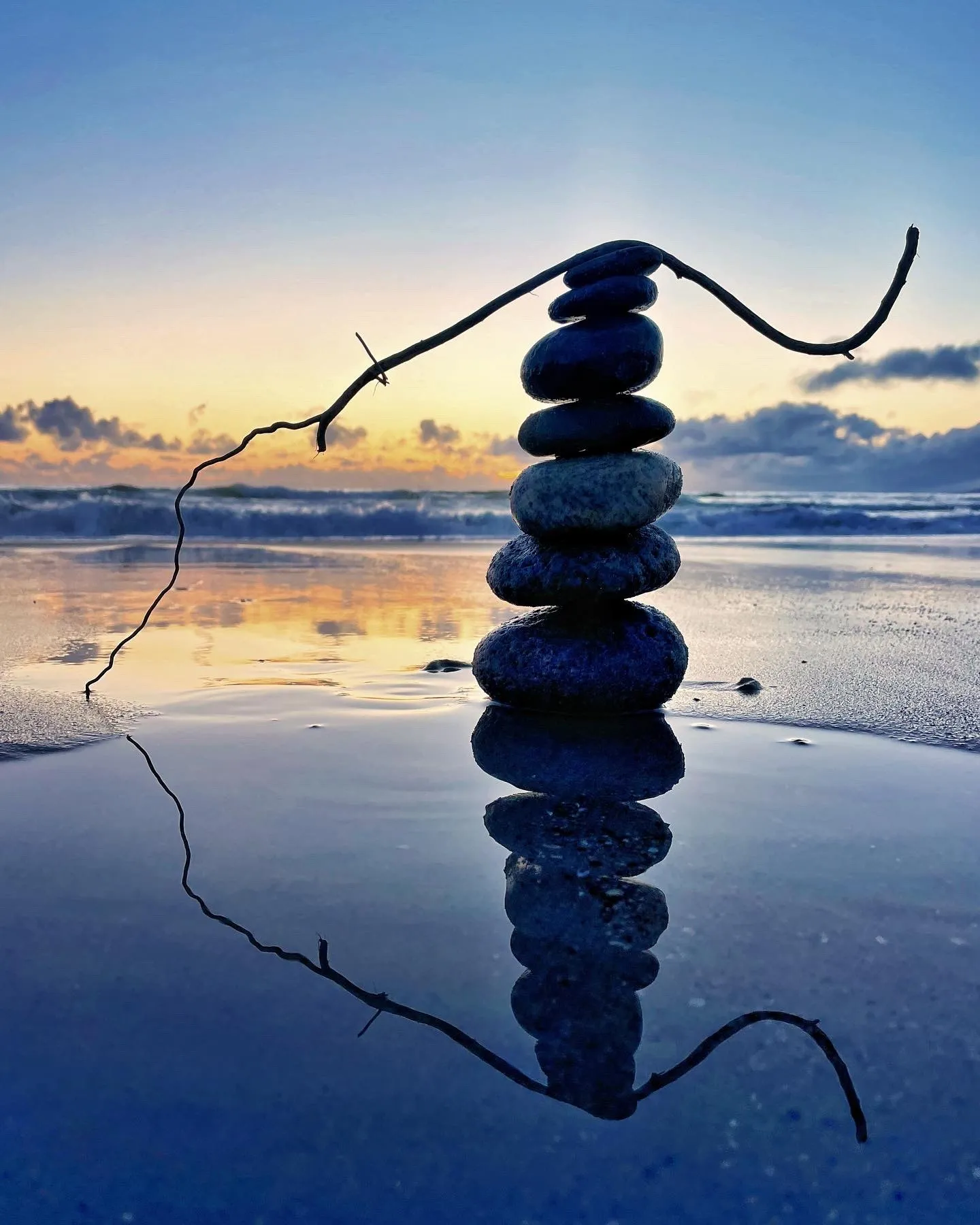 A tall stone stack with branching driftwood mirrored in still water at dusk