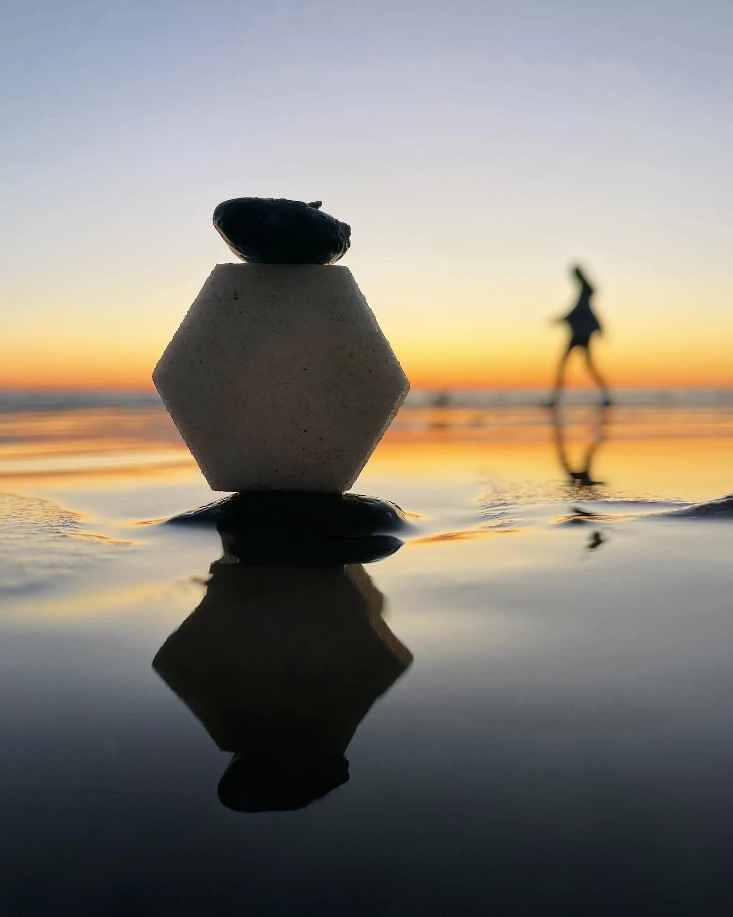 A diamond-shaped stone balanced above its reflection with a distant figure beyond