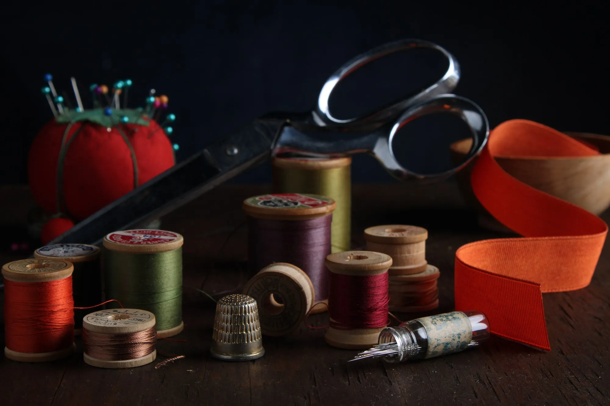 Sewing scissors, thread spools, and red ribbon form a still life on a dark table.