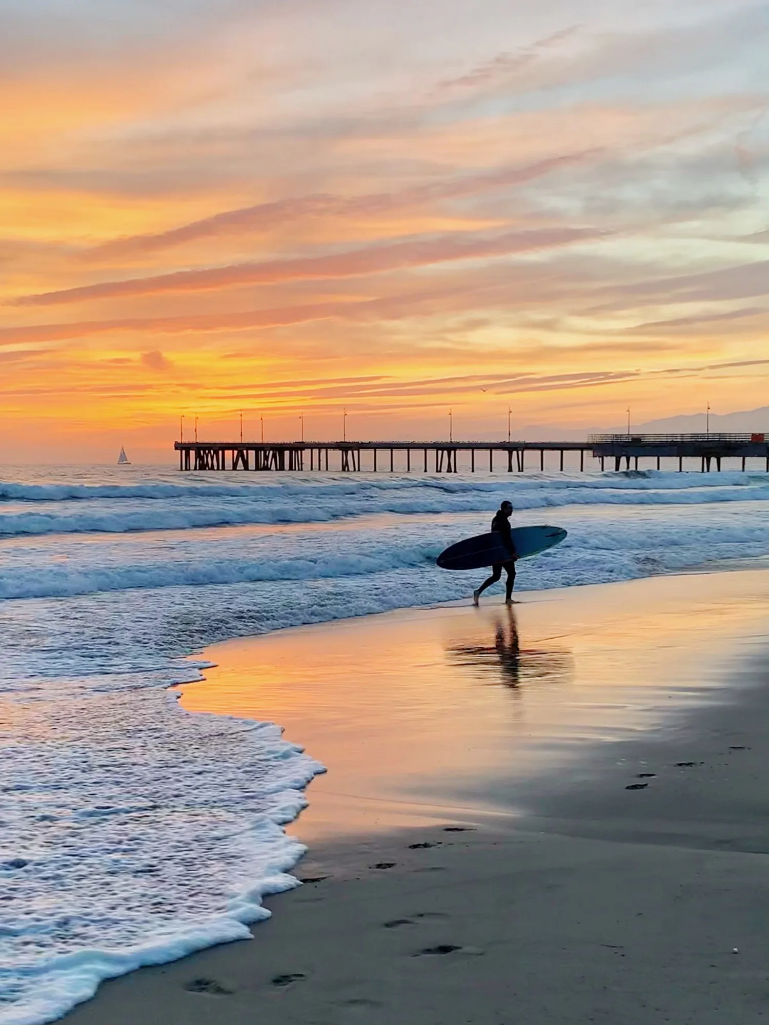 A small surfer walks beside the water under a pale apricot sky.