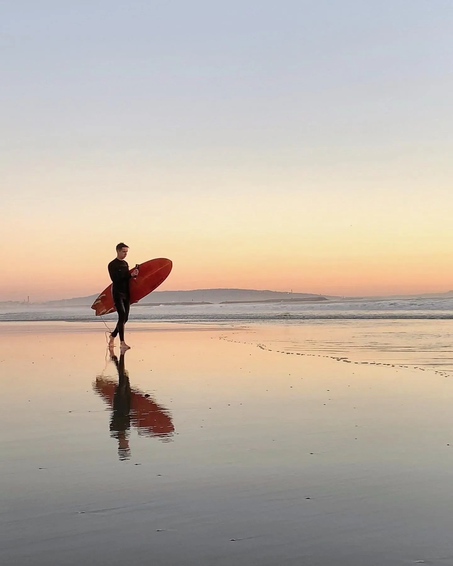 A surfer carries a board across the shallows. A beach observation made in the changing evening light.