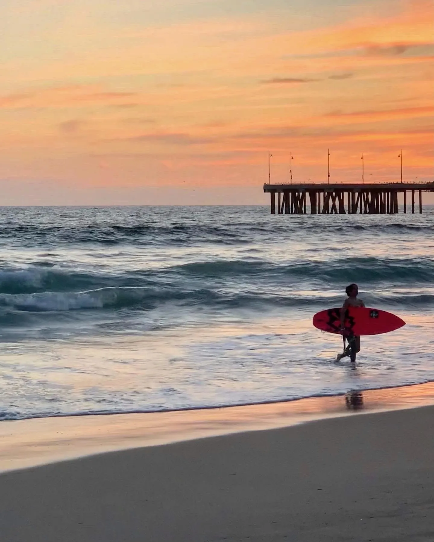 A surfer walks beneath the pier through orange evening light.