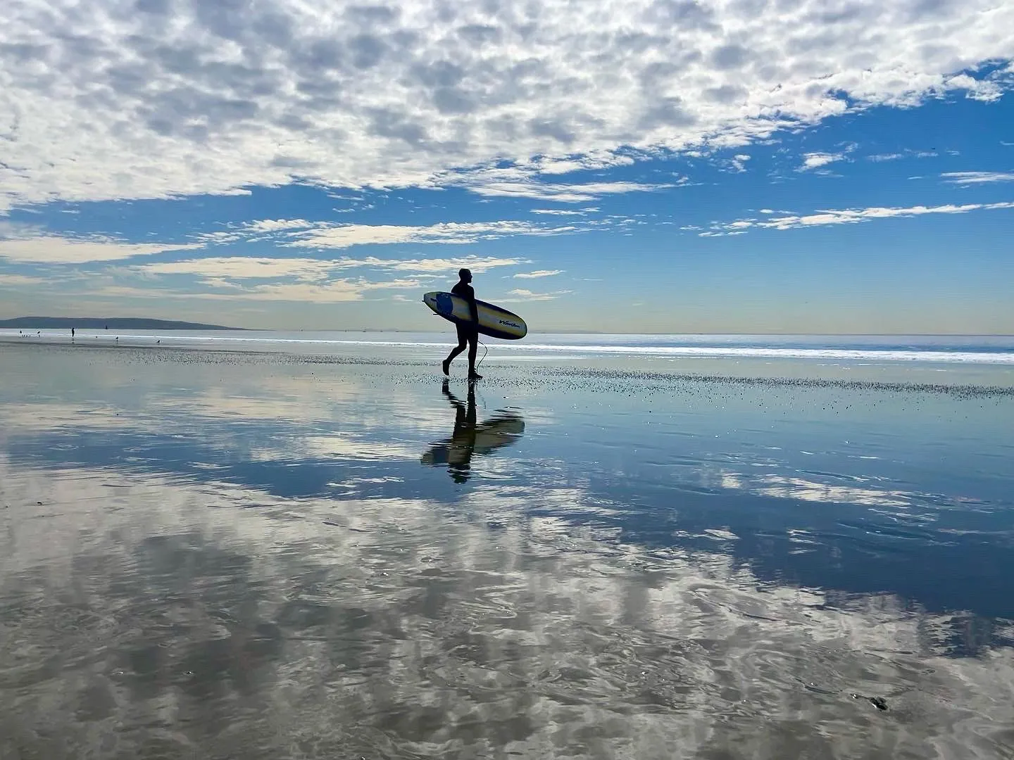 A surfer reflected in wet sand under a wide cloudy sky