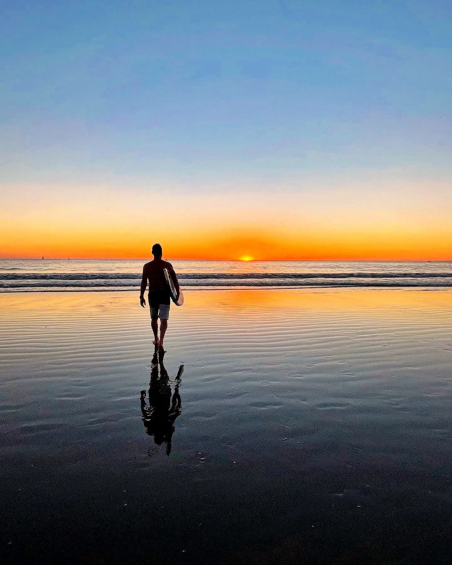 A lone surfer walking toward a narrow band of orange sunset light