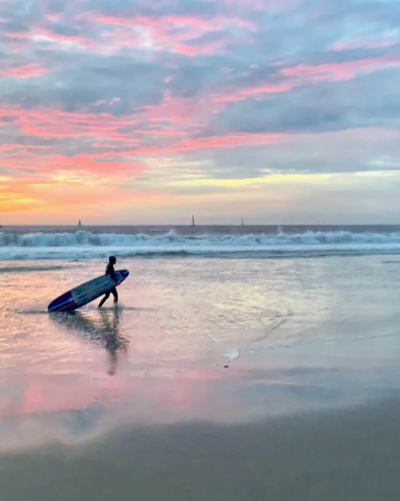 A surfer carrying a blue board beneath a pink and blue clouded sky