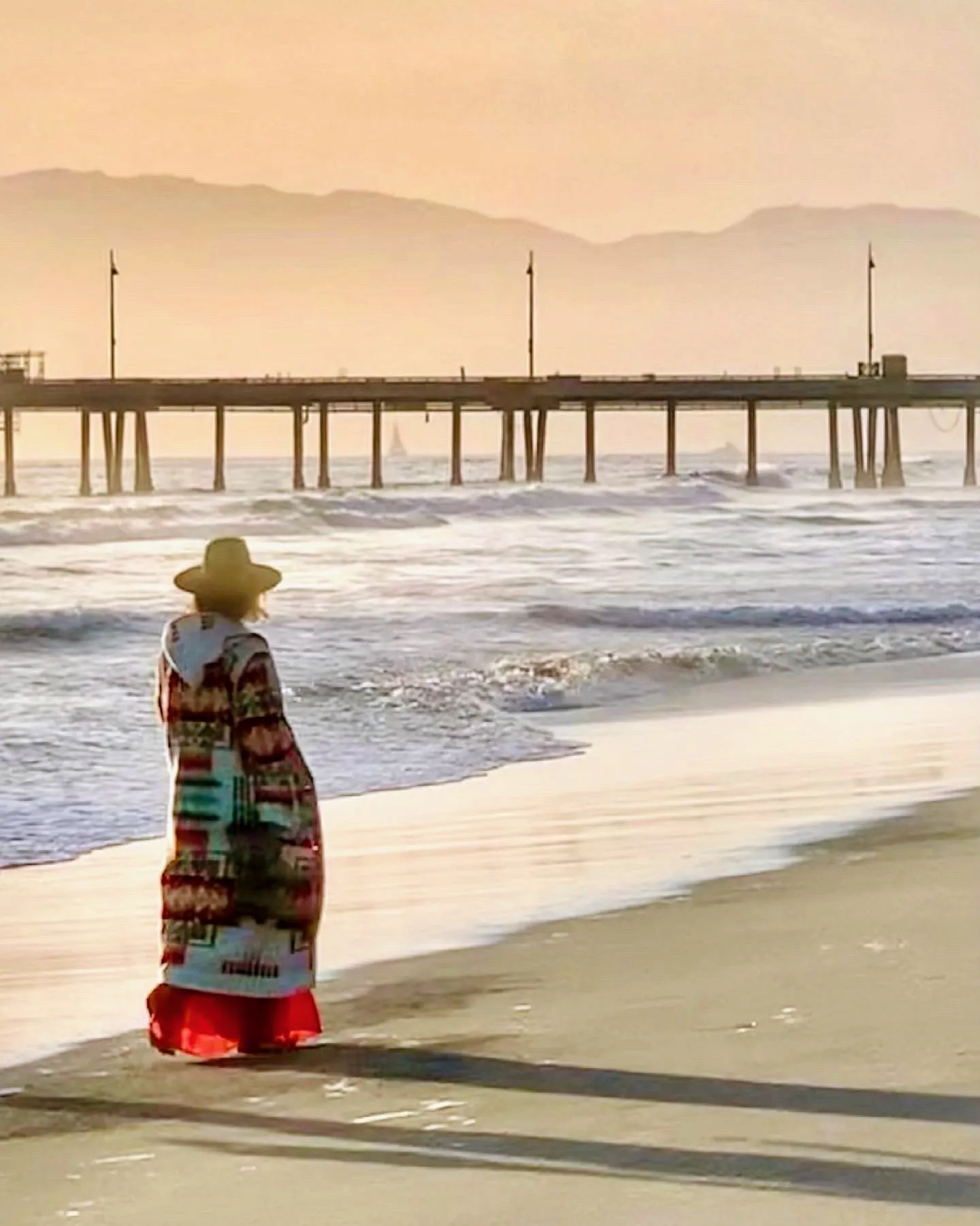 A woman in a patterned dress stands at the shoreline beneath a pastel sky.